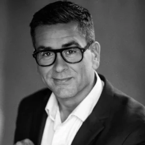 black and white picture of Miles Phillips from Miles marketing Ltd, he is wearing a black jacket, white shirt and black glasses, he is clean shaven